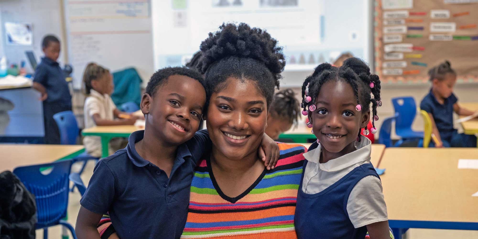 Smiling adult with arms around two young students in a classroom.