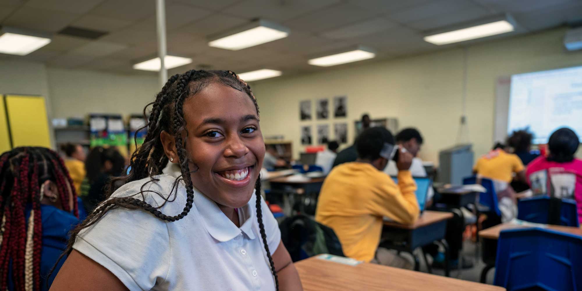 Smiling student with long braids sitting at a desk in a busy classroom.