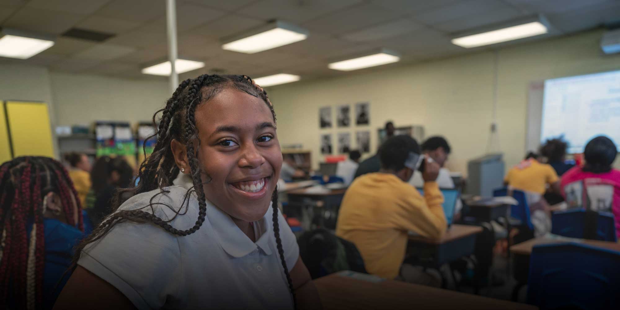 Smiling student with long braids sitting at a desk in a busy classroom.