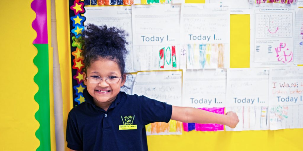 Elementary student smiling and pointing to work on a bulletin board.