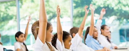 Elementary students with hands raised at desk.